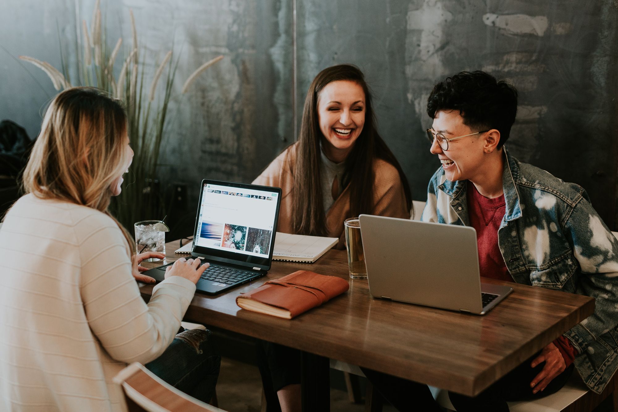 Three colleagues laughing together while working on laptops at a wooden table