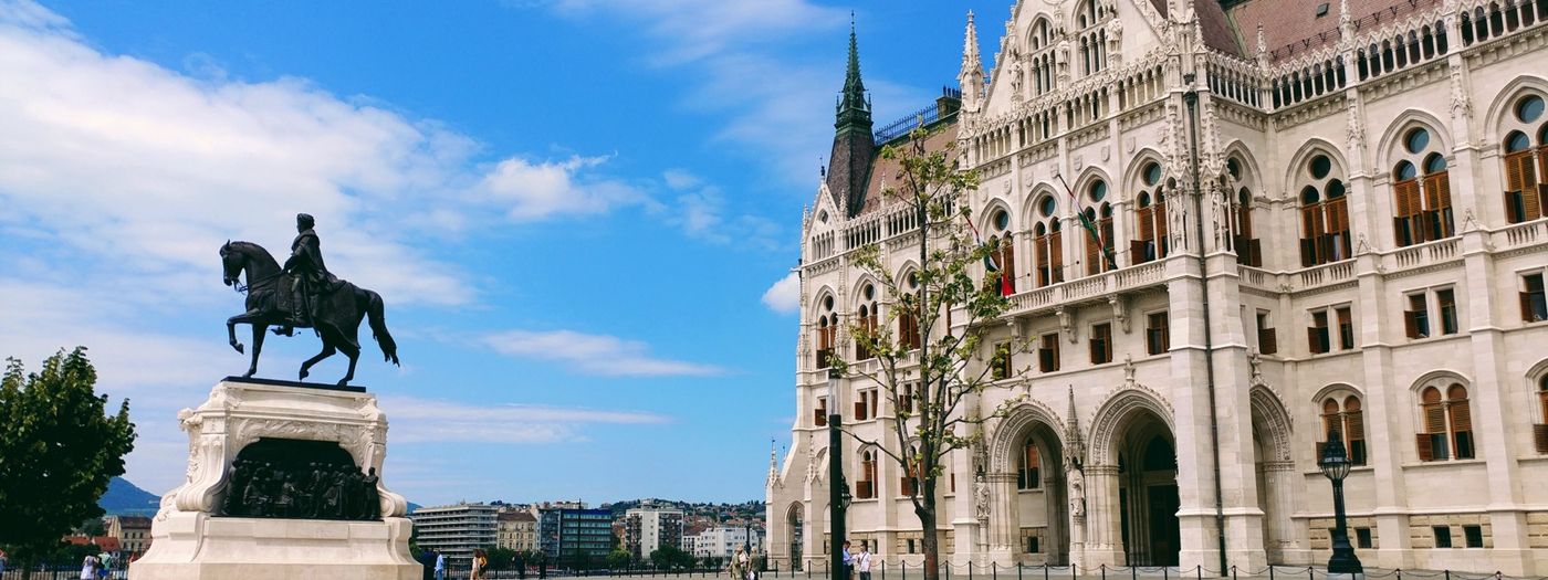 A neo-gothic parliament building beside an equestrian statue under a blue sky